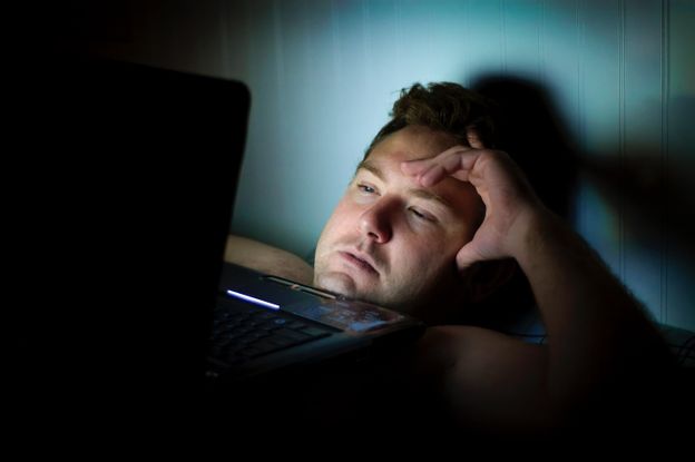 Exhausted man laying in bed with his laptop, working late.